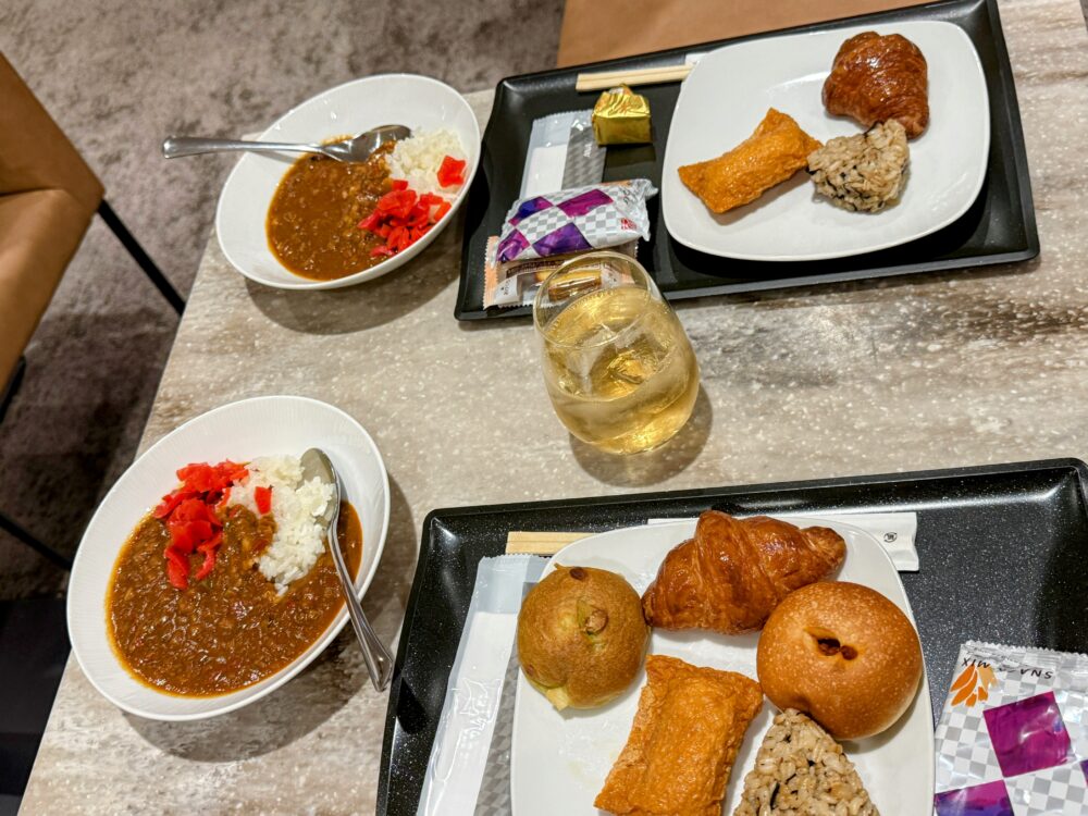 羽田空港ANAスイートラウンジで提供される軽食メニュー。ANAオリジナル野菜カレー、いなり寿司、おにぎり、パン、スナック、ウイスキーのロックなどが並ぶテーブル。