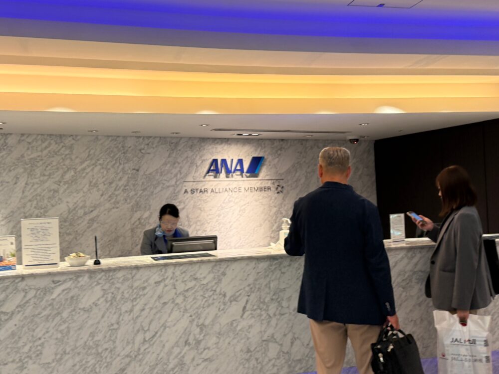 羽田空港ANAスイートラウンジの受付カウンター。大理石調の壁にANAロゴが映える上品な空間で、スタッフが丁寧にチェックイン対応をしている。