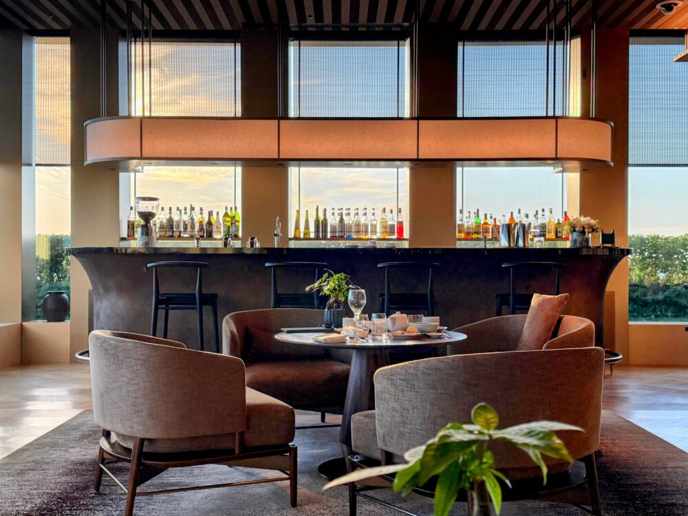 ザ・リッツ・カールトン福岡 クラブラウンジの夕暮れ時のバーエリア。手前の丸テーブルにはアフタヌーンティーのセットが置かれ、奥には特徴的なデザインのバーカウンターと、窓から見える夕景が広がっている。