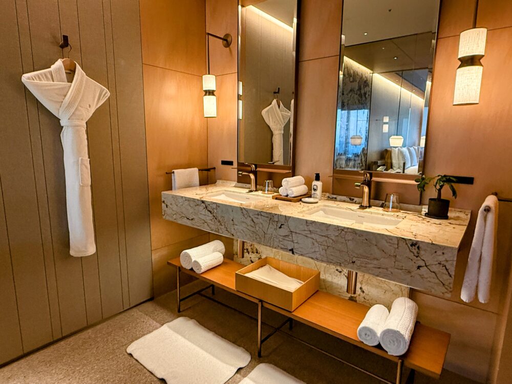 大理石の壁とダブルシンクが特徴的なリッツ・カールトン福岡の洗面台エリア。厚手のバスローブが掛けられており、鏡にはベッドルーム側の景色が映り込んでいる。