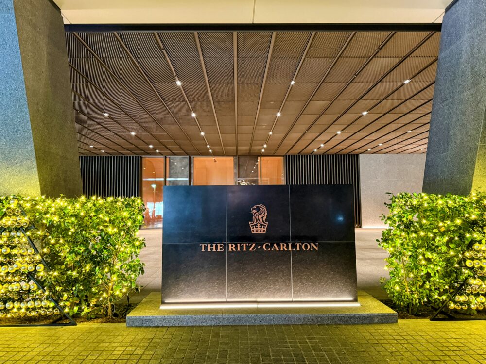 リッツ・カールトン福岡の正面玄関。黒い石造りのサインボードに金色のライオンロゴとホテル名が輝き、両脇にはクリスマスイルミネーションで飾られた植栽がある。