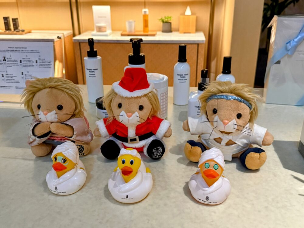 リッツ・カールトン福岡のスパ受付に並ぶ、サンタクロース衣装や和装のリッツ・ライオンのぬいぐるみと、頭にタオルを巻いてキュウリパックをしたユニークなラバーダックたち