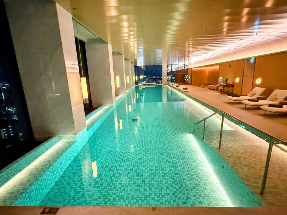 リッツ・カールトン福岡のインドアプール（夜）。水底からのライトアップで水面がエメラルドグリーンに輝き、窓の外には夜景が広がっている