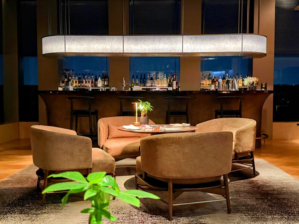 24階クラブラウンジの窓側席から望む博多湾の夕景、バーカウンターが柔らかく点灯（カクテルタイム）