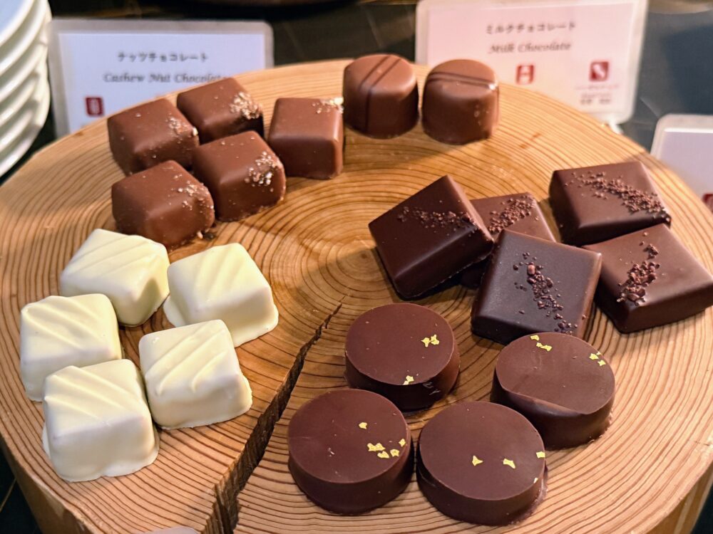 木の切り株のプレートに並べられた、種類豊富なボンボンショコラ（ビター、ミルク、ホワイトなど）。