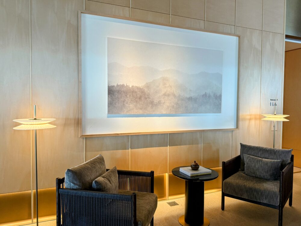 明るい木目の壁に飾られた、霧のかかった山並みを描いた大きなアート作品と、その手前にあるモダンなグレーのチェアとサイドテーブルの休憩スペース