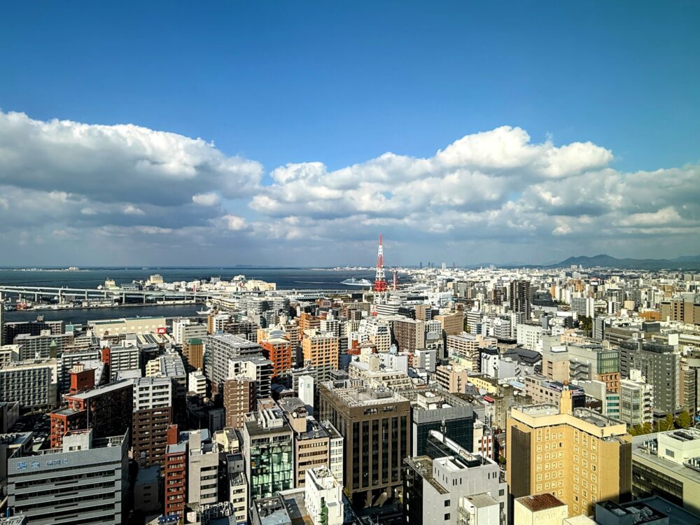リッツ・カールトン福岡の客室から望む昼間の博多の街並み。青空の下、博多湾と赤い博多ポートタワーが鮮やかに見える