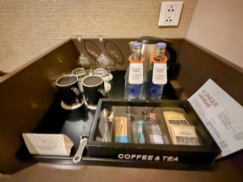 ミニバー上のトレイにセットされた無料の飲料水2本、マグカップ、グラス。「COFFEE & TEA」と書かれた木箱の中にティーバッグや砂糖が収納されている。