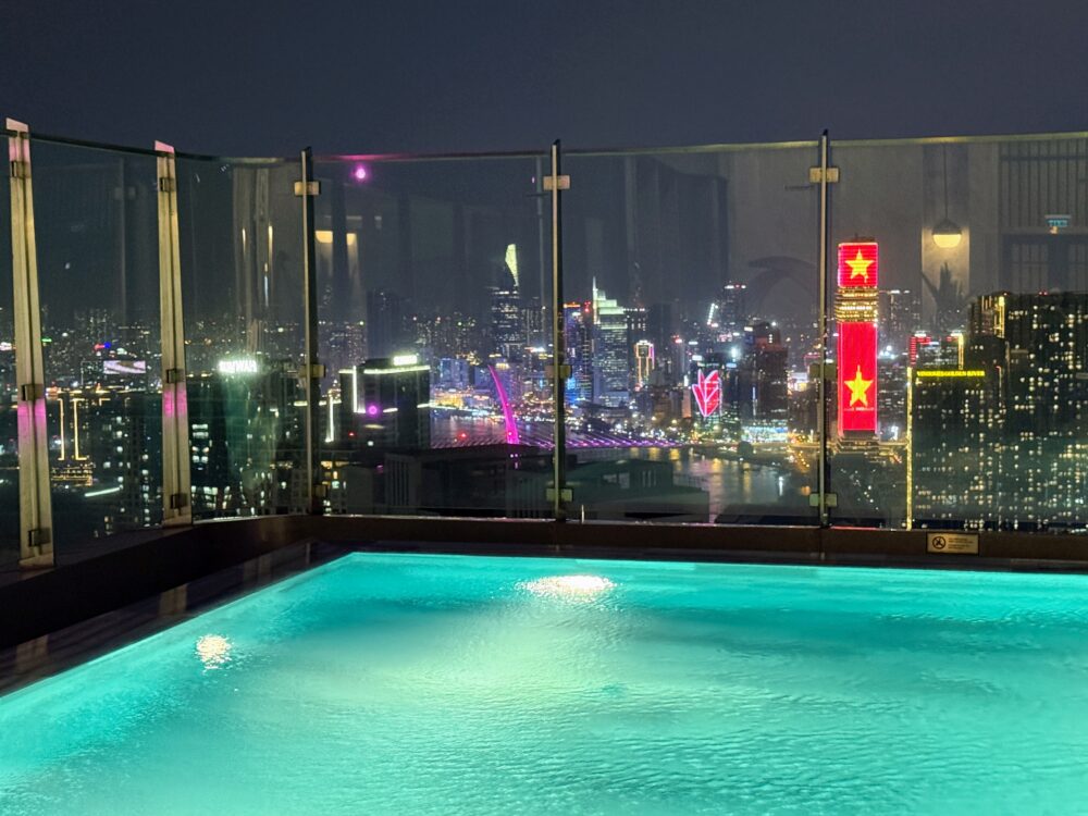鮮やかなターコイズブルーに光る夜のプール水面。背景には赤くライトアップされたビルや、きらめく街のイルミネーションが見える。