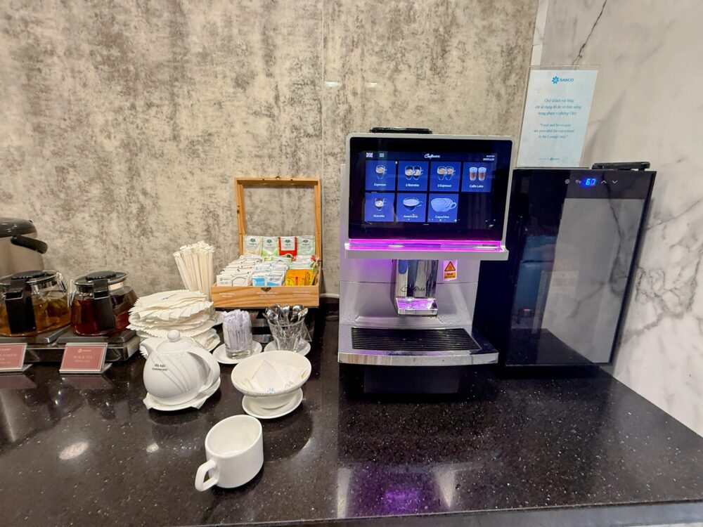 タッチパネル式の全自動エスプレッソマシン（Cafematic）。画面にはエスプレッソ、カプチーノ、カフェラテなどのメニューアイコンが表示されている。左側にはティーバッグが入った木箱、右側にはミルククーラーが設置されている。