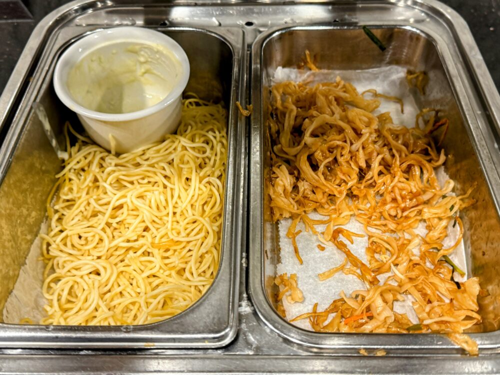 ビュッフェ台に並ぶ2種類の麺料理。左側のバットにはプレーンなスパゲッティ麺が、右側のバットには茶色く炒められた焼きそばのような麺が大量に盛られている。奥には麺にかけるための白いクリームソースが入った容器がある。