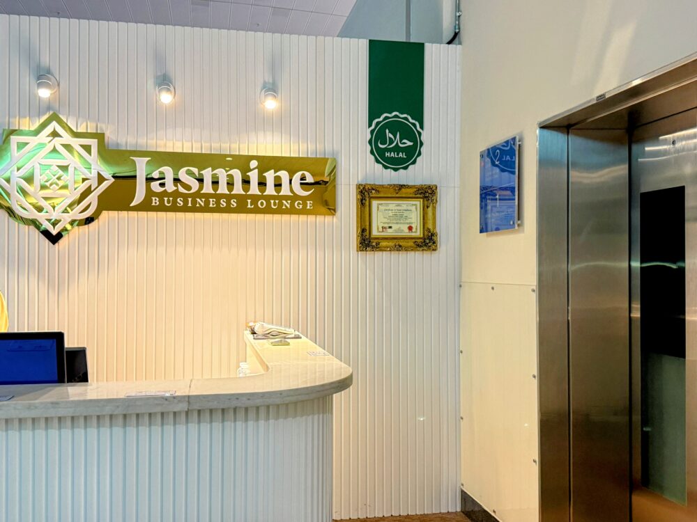 ラウンジ内部の受付カウンター。白を基調とした清潔感のあるデザインで、壁面には金色の「Jasmine」のロゴと緑色のハラル認証マークが掲示され、右手に下階へ降りるエレベーターがある。