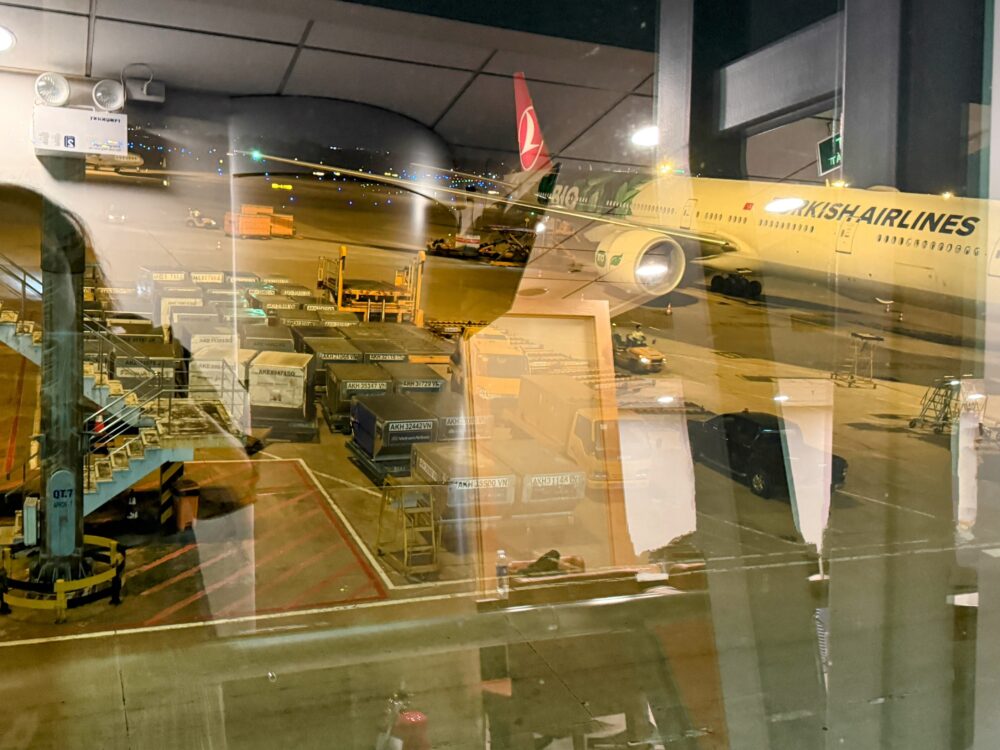 ラウンジの窓から見える夜の駐機場の様子。ガラス越しに、ターキッシュ・エアラインズ（Turkish Airlines）の大型機がすぐ目の前に駐機しているのが見える。手前には貨物コンテナや作業車両が並んでいる。