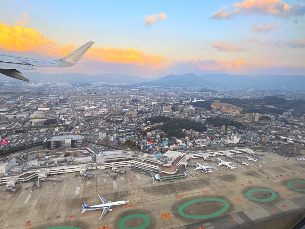 福岡空港を離陸した飛行機の窓から見た景色。滑走路と並ぶANA機が見え、遠くには福岡市街と山々、夕焼けに染まる雲が広がっている。