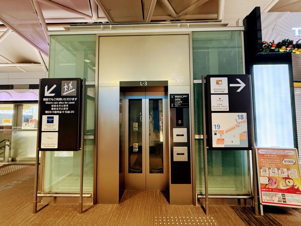 中部国際空港（セントレア）国際線制限エリア内、19番ゲート付近にあるラウンジ行きエレベーターの入口。左側の案内板には「The Coral Lounge（ザ・コーラル・ラウンジ）」のロゴがあり、エレベーターのフロア案内表示には「2F ラウンジ」と記されている。