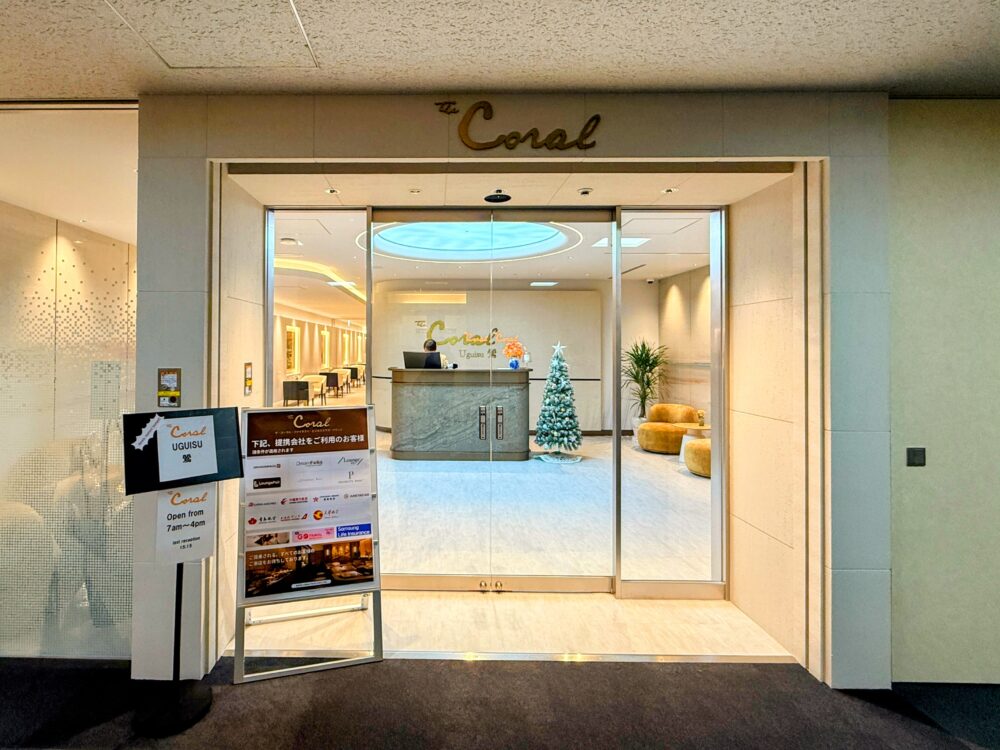 中部国際空港コーラルラウンジ鶯（うぐいす）の正面入口。ガラス扉の奥に石造りの受付カウンターとクリスマスツリーが見える。左手には営業時間（7:00-16:00）やプライオリティパス等の提携案内ボードが設置されている。