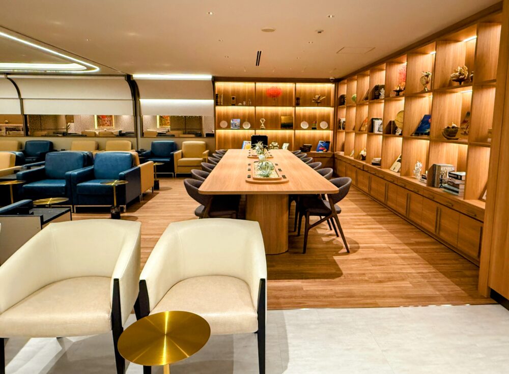 コーラルラウンジ鶯（うぐいす）のメインエリア。手前にはゆったりとした白いアームチェアとゴールドのサイドテーブル、中央には植物が飾られた長い木製テーブルがあり、奥の壁一面には本やオブジェが並ぶ木棚がライトアップされている。温かみのあるライブラリーのようなリラックス空間。