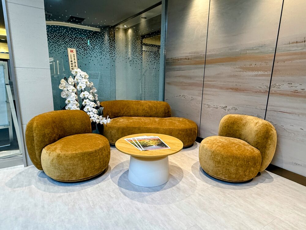 コーラルラウンジ鶯（うぐいす）にある、マスタードイエローの丸みを帯びたユニークなベルベット調ソファー。白い胡蝶蘭と抽象的なアートが飾られた、フォトジェニックなコーナー。