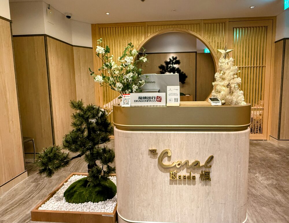 中部国際空港コーラルラウンジ楓（かえで）の受付。手入れされた松の盆栽と円窓のデザインが和の雰囲気を演出している。カウンターには「The Coral Kaede 楓」のゴールドのロゴがある。