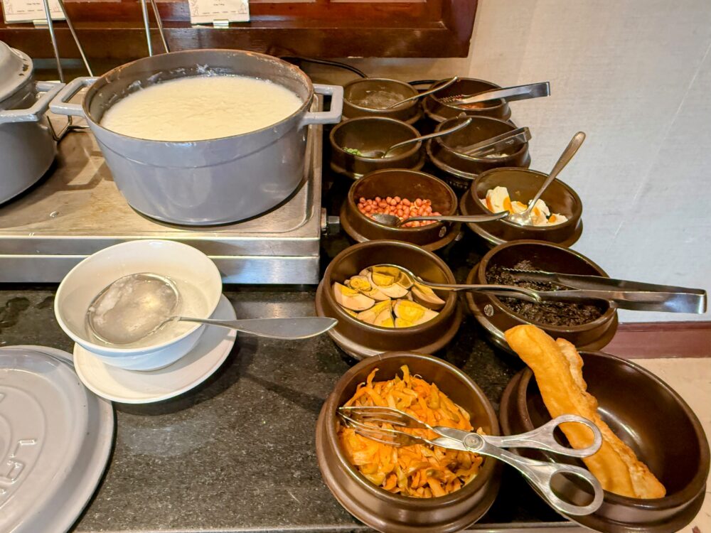 大きな鍋に入った中華粥(Congee)と、ピータンや揚げパンなどの豊富なトッピング類。