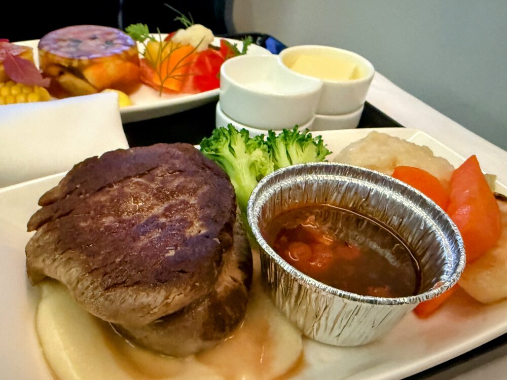 ANAビジネスクラス洋食コースのメインディッシュ。厚みのある牛フィレ肉のステーキに、別添えのソース、マッシュポテト、ブロッコリーなどの温野菜が添えられている。奥には彩り豊かな前菜プレートも見える。