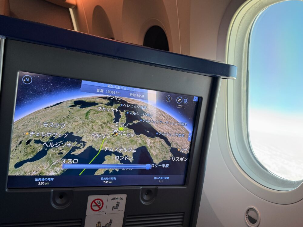 機内モニターに表示されたフライトマップ。ヨーロッパ上空を飛行中の現在地と、窓から見える青空。