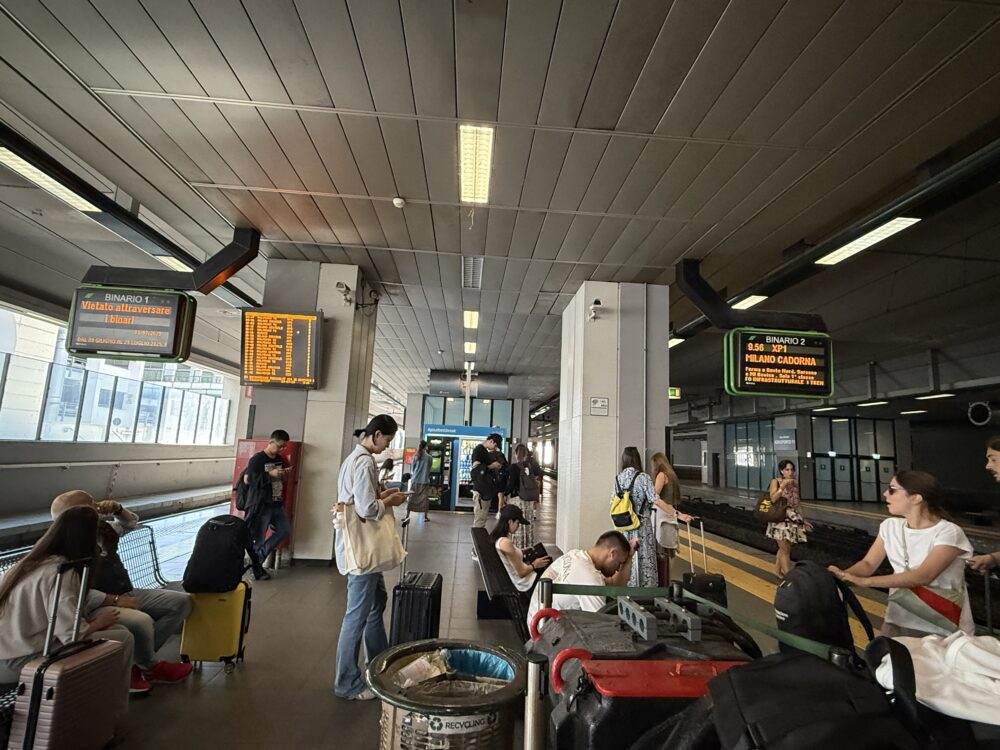 マルペンサ空港駅のホームで電車の到着を待つ様子。電光掲示板には「MILANO CADORNA(ミラノ・カドルナ)」行きの表示がある。