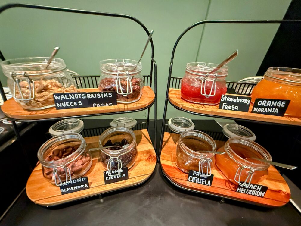 木製のスタンドに並べられたガラス瓶入りのジャム(イチゴ、オレンジ、ピーチ)と、クルミやレーズンなどのドライフルーツ・ナッツ類。