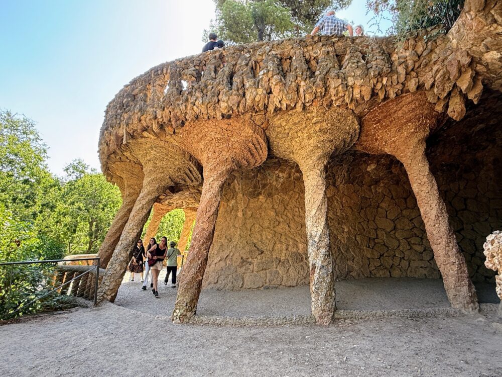 グエル公園の高架歩道(ヴィアドゥクト)。ガウディ設計の石造アーチと木の幹のような柱が特徴で、下の通路を人々が歩いている。