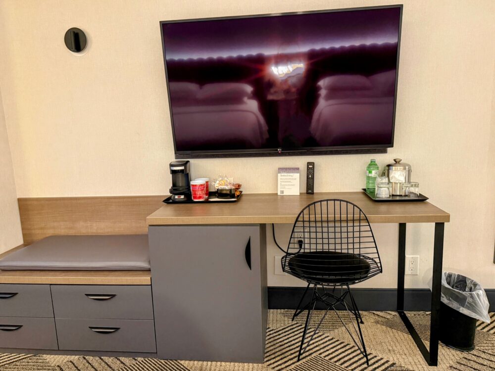 壁掛けの大型テレビと、作業しやすい広さのデスク。デスク下には冷蔵庫が収納されており、ワークチェアも完備。