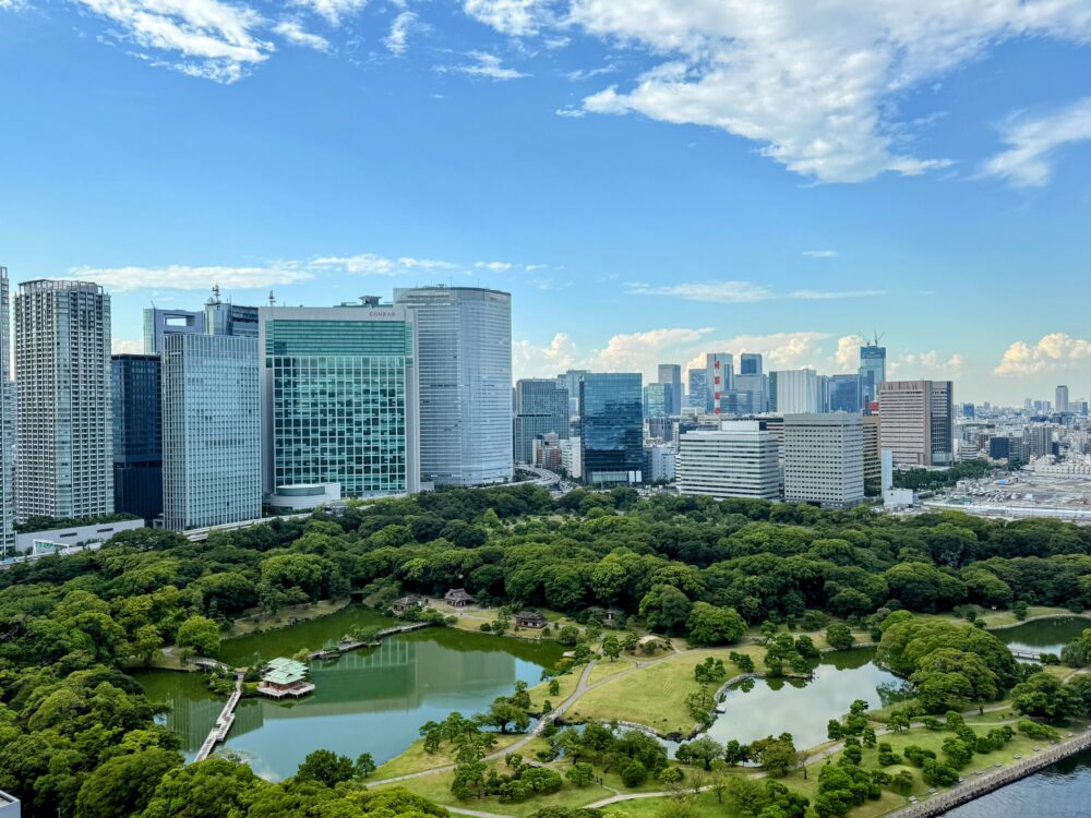 メズム東京の客室から見下ろす、広大な浜離宮恩賜庭園の全景。手前には豊かな緑と池、茶屋が広がり、背景にはコンラッド東京などの汐留の高層ビル群と青い空が対比的に映し出されている。