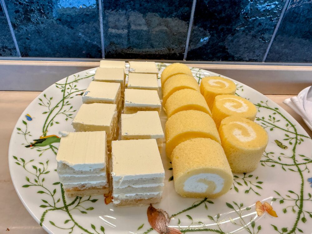 花柄の丸皿に並ぶ2種類のケーキ。左側は白いムース層が美しいガトーブランのカット済みのもの、右側はクリームを巻いたロールケーキのスライスが整然と並んでいる。手前にホテルのメニュープレートが置かれている。