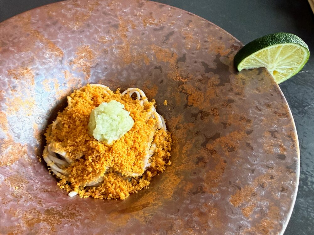 錆色のアンティーク風の丸皿に盛られた唐墨蕎麦。細い蕎麦の上にオレンジ色の唐墨がたっぷりとかかり、わさびが添えられている。皿の端にライムが一切れ置かれている。