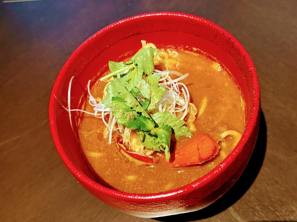 赤い漆器風のボウルに盛り付けられたオマール海老とトリュフのラーメン。オレンジ色の濃厚なスープにオマール海老の爪と麺が入り、青ネギとパクチーが添えられている。
