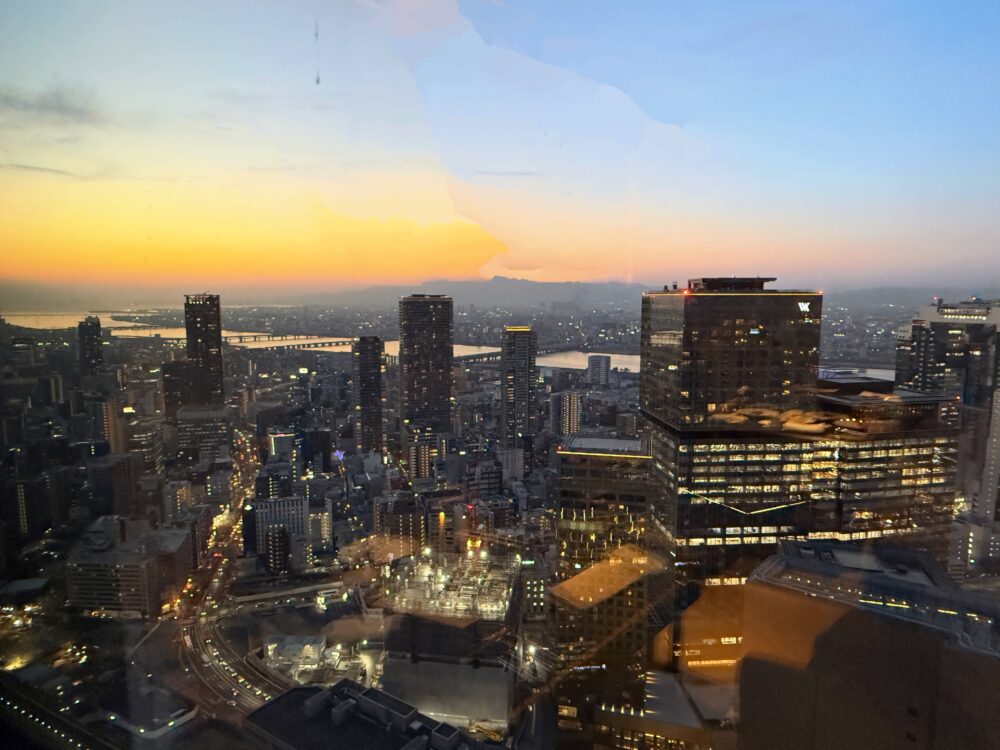 スペシャリティサロンから望む大阪の夕景。オレンジとブルーのグラデーションに染まる空の下、大阪の街並みと淀川・大阪湾方面が一望できる。街のあちこちに灯りがともり始め、昼から夜へと移り変わる瞬間が切り取られている。
