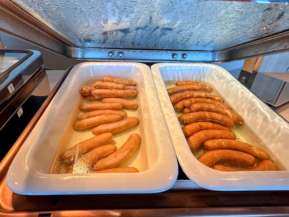 大阪ステーションホテル スペシャリティサロンの朝食ビュッフェのホットミールコーナー。2種類のソーセージが白いトレイに並ぶ。