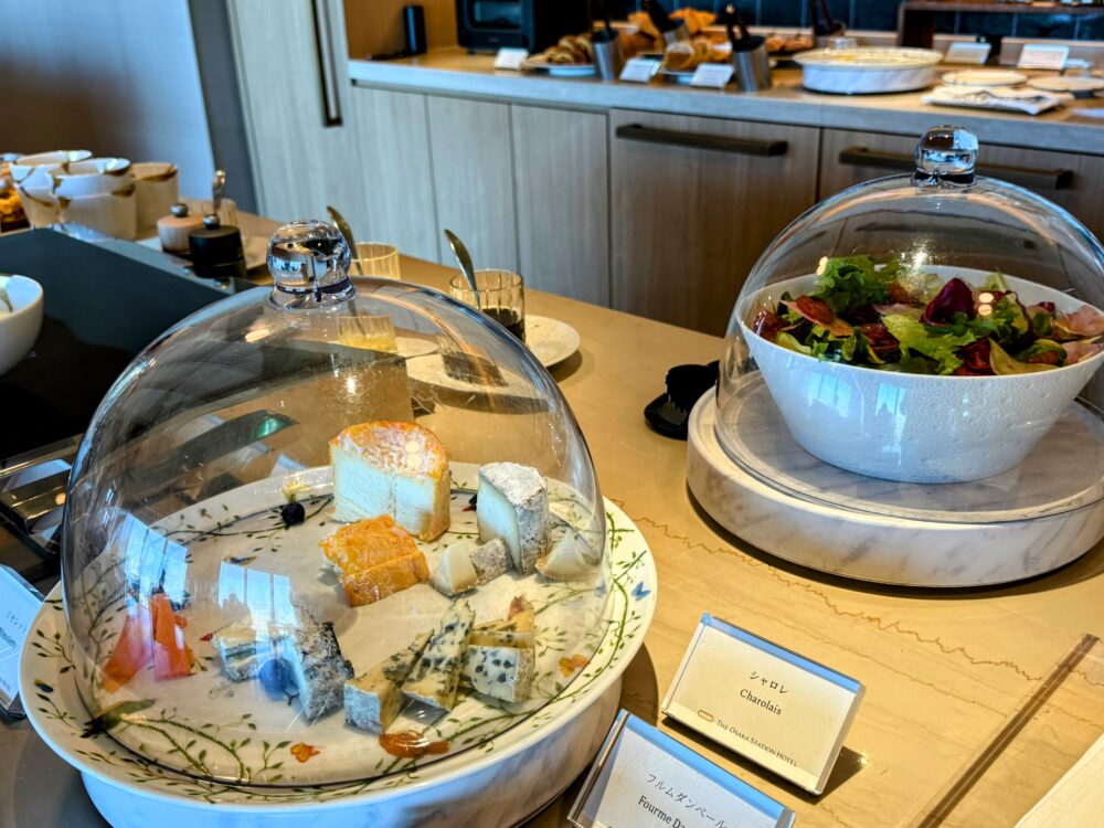 大阪ステーションホテル スペシャリティサロンの朝食ビュッフェのチーズコーナー。シャロレ・フルムダンベール等のフランス産チーズがガラスドームに入って提供され、隣にはサラダも並ぶ。