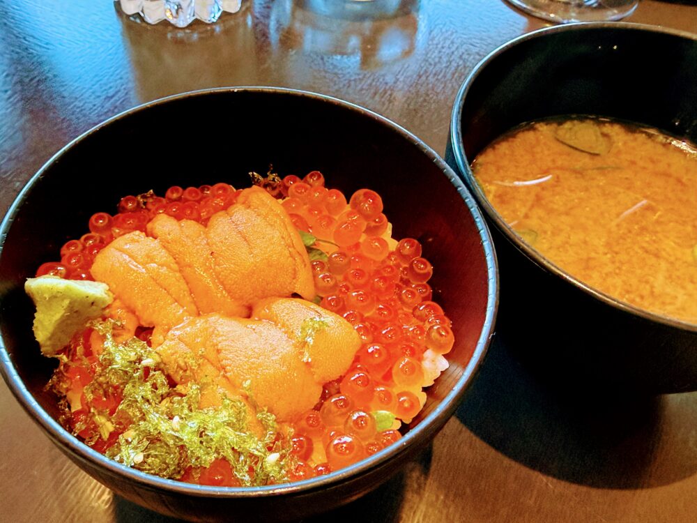 黒い丼ぶりに盛られた雲丹といくらの丼ぶりのアップ。いくらがたっぷりと盛られた上に雲丹が並び、青のりとわさびが添えられている。右側には赤みそのしじみ汁が見える。