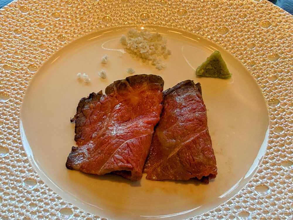 白い丸皿に盛られた黒毛和牛のロティ。薄くスライスされた牛肉2枚が重なり、わさびと塩が添えられている。