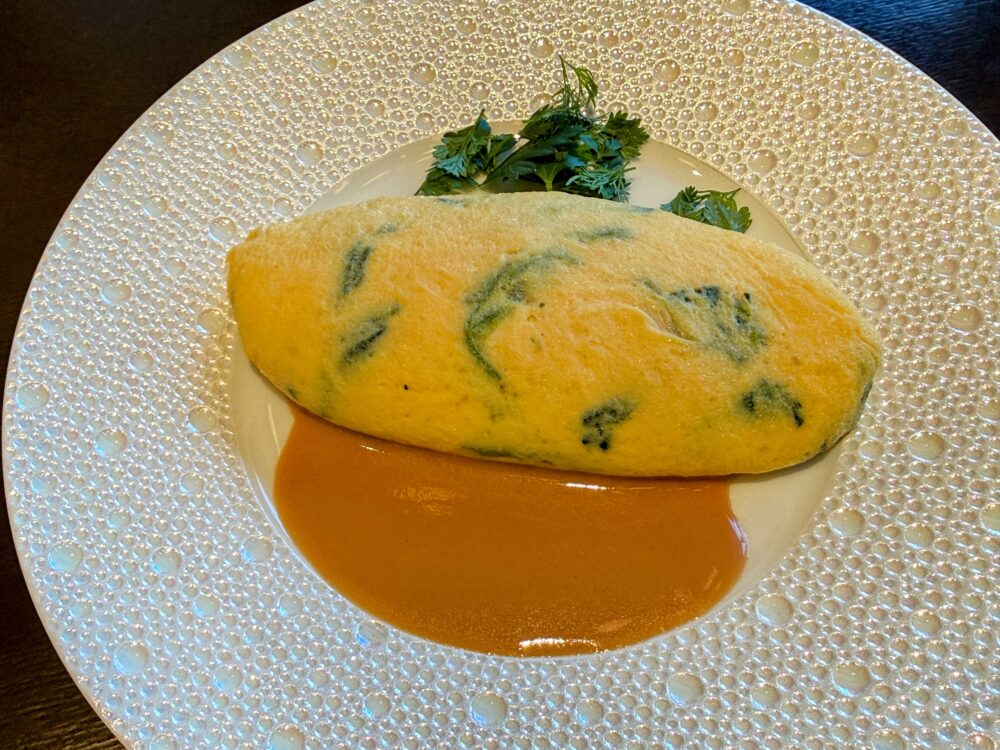 白い丸皿に盛られたほうれん草とチーズのオムレツ。ほうれん草が生地に混ぜ込まれた緑がかったオムレツの下にビスクソースが広がり、上にパセリとディルが添えられている。