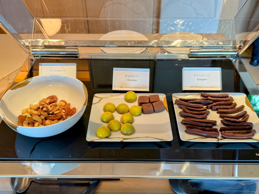 ガラスケース内に並ぶミックスナッツ・チョコレート・オランジェット。左の白いボウルにミックスナッツ、中央の皿に抹茶色のボンボンショコラとチョコレート、右の皿にオランジェットが盛られている。