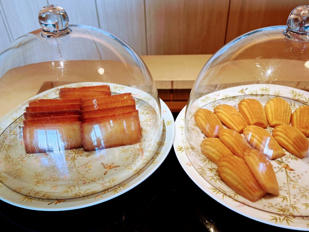 ガラスドームで覆われた2種類の焼き菓子。左の皿にキャラメル色のフィナンシェ、右の皿に貝殻型のマドレーヌが花柄の皿に整然と並んでいる。