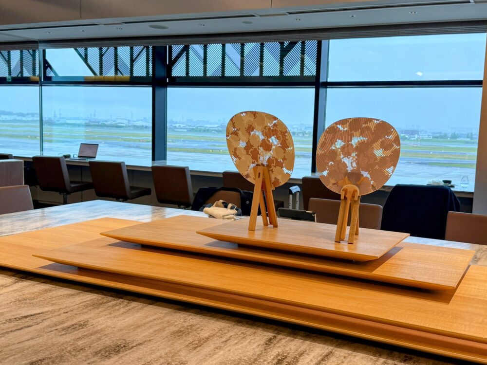 伊丹空港ANAスイートラウンジの工芸品テーブルに展示された、阿以波の京うちわ2点。雪輪文様が施された丸型のうちわが木製スタンドに立てられ、背景には滑走路が広がる。