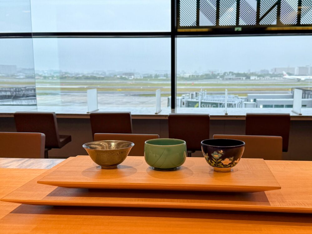 伊丹空港ANAスイートラウンジの工芸品テーブルに並ぶ、西川貞三郎商店の京焼・清水焼3点。金彩・青磁・黒地花柄とそれぞれ異なる表情を持つ茶碗が、滑走路を望む窓の前に展示されている。