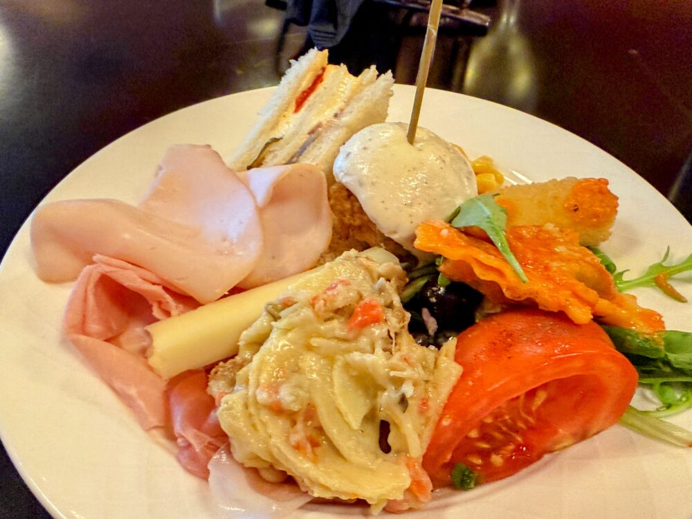 ラウンジでの軽食プレート。ピックが刺さったクラブサンドイッチ、たっぷりのハム、トマト、クリーミーなサラダの盛り合わせ。