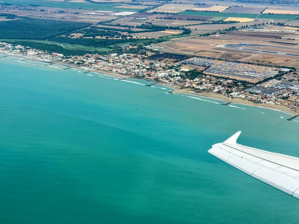 飛行機の窓から見下ろすローマ・フィウミチーノ空港周辺の美しい海岸線。鮮やかなターコイズブルーの海と白い砂浜、緑豊かな大地の上を飛ぶ飛行機の翼。