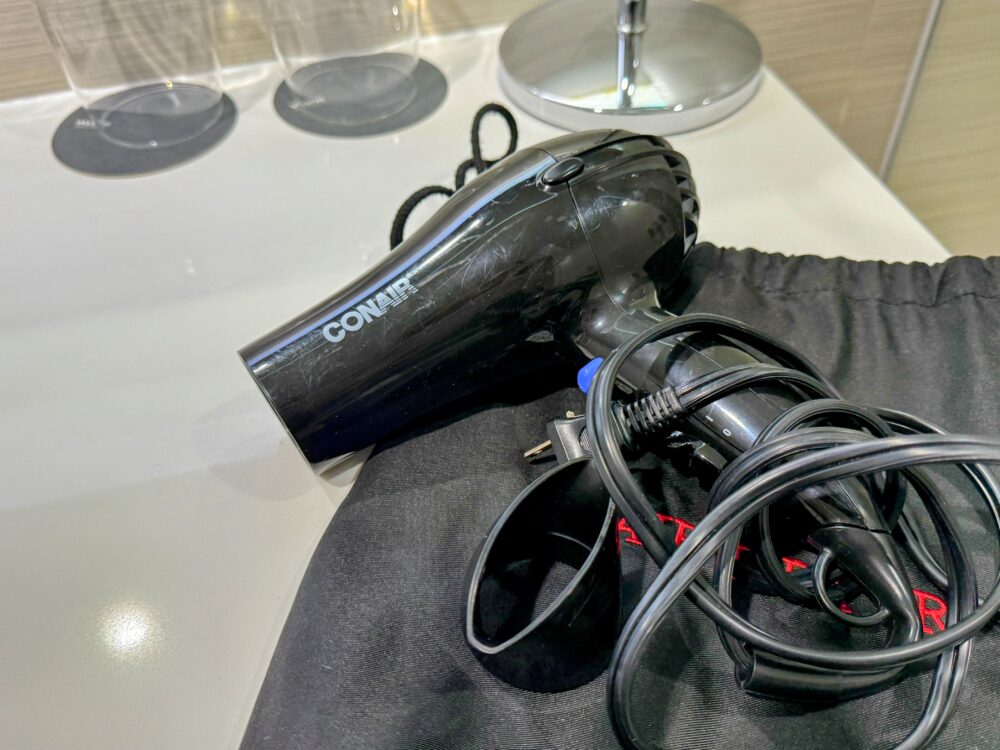 黒い巾着袋の上に置かれたCONAIR製の黒いヘアドライヤー。コードが束ねられている。
