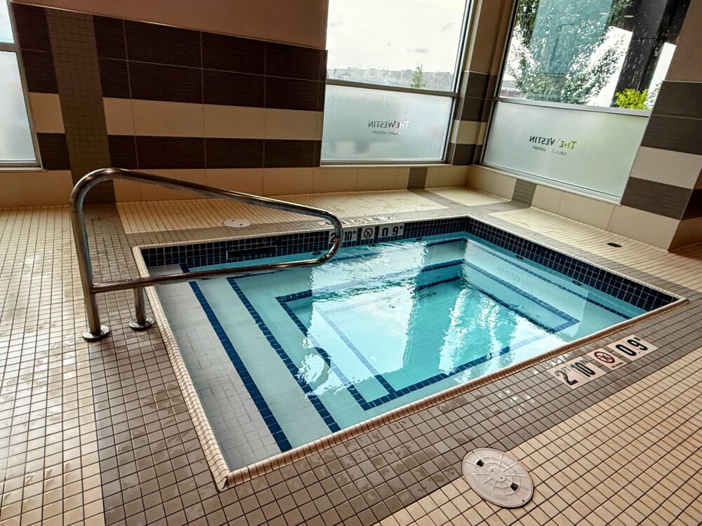 タイル張りの四角い屋内ジャグジー(ホットタブ)。手すり付きの階段があり、窓には「THE WESTIN CALGARY AIRPORT」の文字が見える。