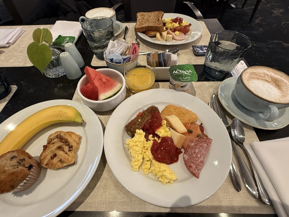 実際に選んだ朝食のテーブル。スクランブルエッグとソーセージのプレート、フルーツの小鉢、バナナとマフィン、そしてカプチーノが並ぶ。