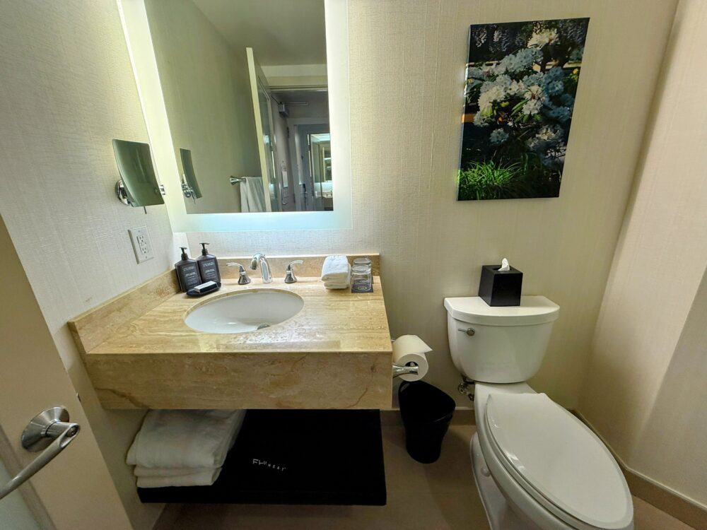 明るいベージュの大理石調タイルで統一されたバスルーム。洗面台、大きな鏡、トイレがあり、壁には花のアートが飾られている。